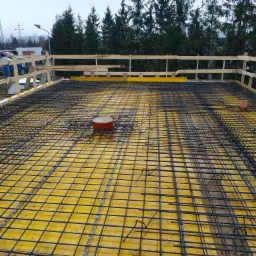 Zbrojenie płyty fundamentowej na żółtym szalunku, widok z góry, z elementami drewnianej konstrukcji zabezpieczającej na krawędzi, w tle drzewa i budynki gospodarcze.