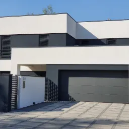 Nowoczesny dom murowany z płaskim dachem, szarą bramą garażową i ogrodzeniem w kolorach bieli i grafitu, widok na elewację frontową i podjazd z kostki brukowej.