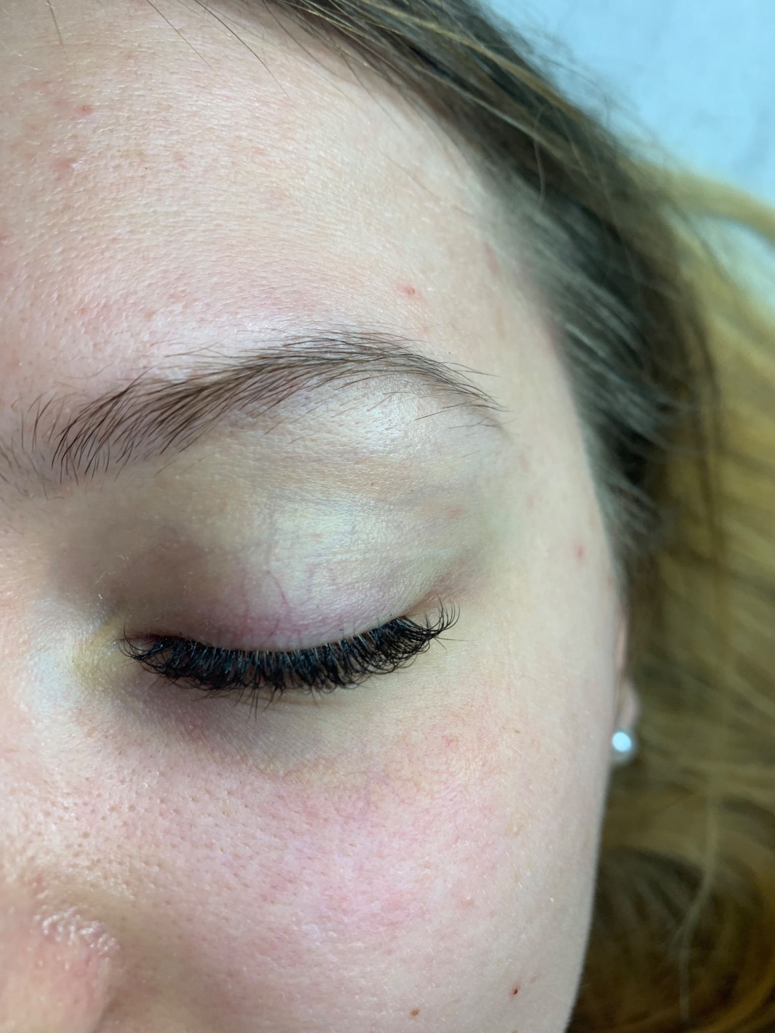 Zbliżenie na zamknięte oko kobiety z przedłużonymi, gęstymi rzęsami; widoczny delikatny makijaż i kolczyk w uchu, skóra z widocznymi porami.