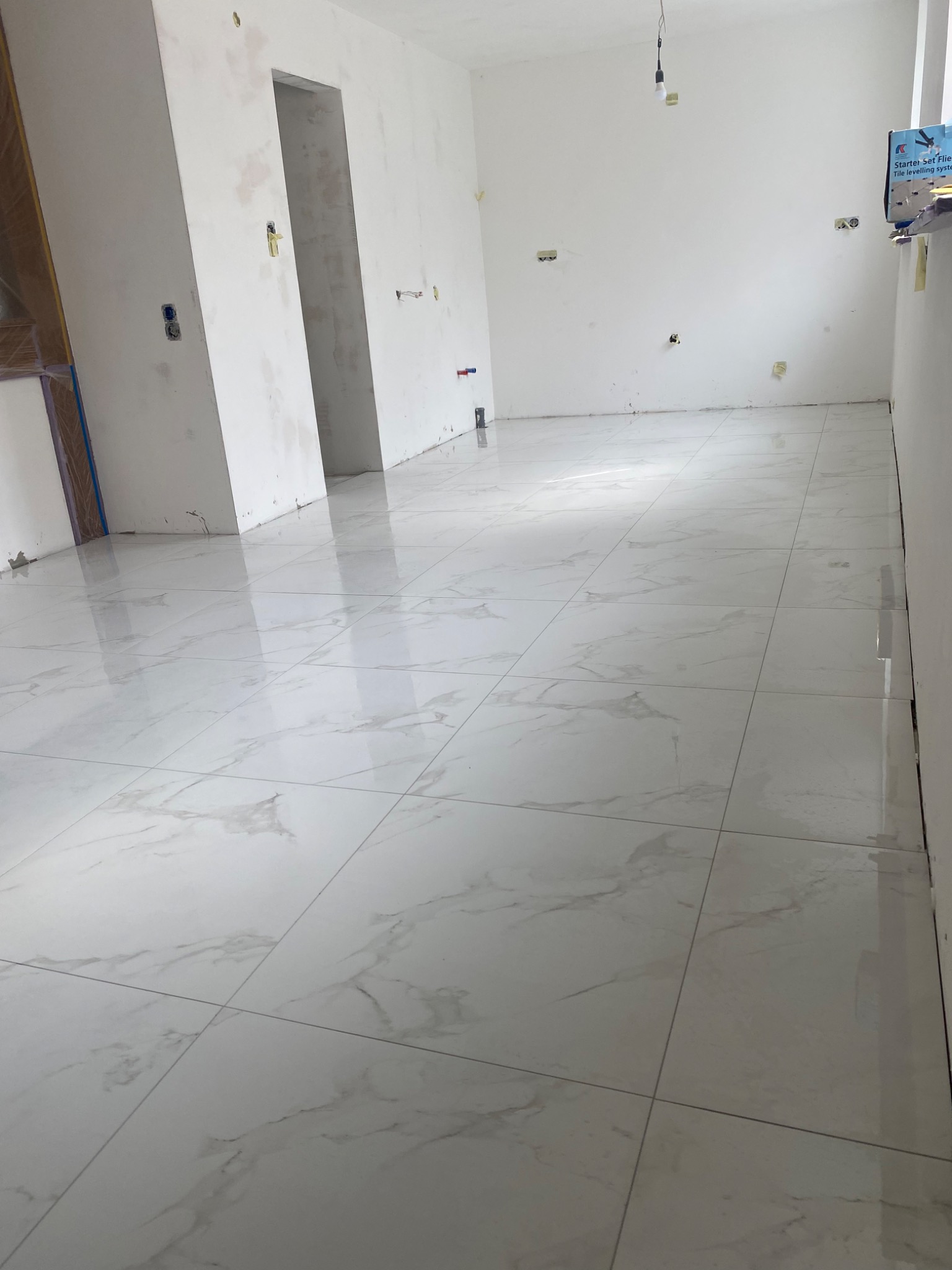 Białe płytki podłogowe z marmurkowym wzorem w surowym wnętrzu, widoczne ślady po instalacjach elektrycznych na ścianach, przygotowane do dalszych prac wykończeniowych.