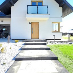 Galanthus - usługi ogrodnicze Piotr Kłosowski - Nowoczesny dom jednorodzinny z wejściem wyłożonym jasnoszarą kostką brukową z ciemnoszarym obramowaniem, otoczony białym żwirem i sztuczną trawą.