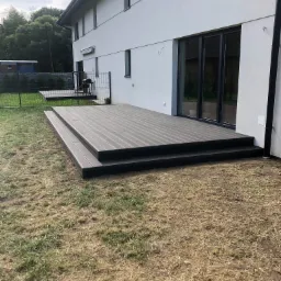 Nowoczesny taras kompozytowy z dwoma stopniami, przylegający do białego budynku z dużymi oknami przesuwnymi, widok z poziomu trawnika.