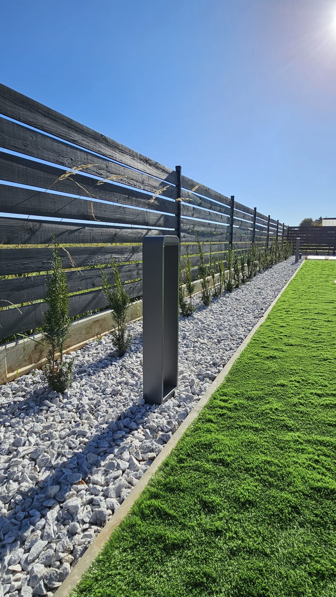 Nowoczesne ogrodzenie z ciemnego drewna z poziomymi szczeblami, oświetlenie ogrodowe w kształcie prostopadłościanu, biały żwir i równo przystrzyżony trawnik, tworzące minimalistyczny krajobraz.