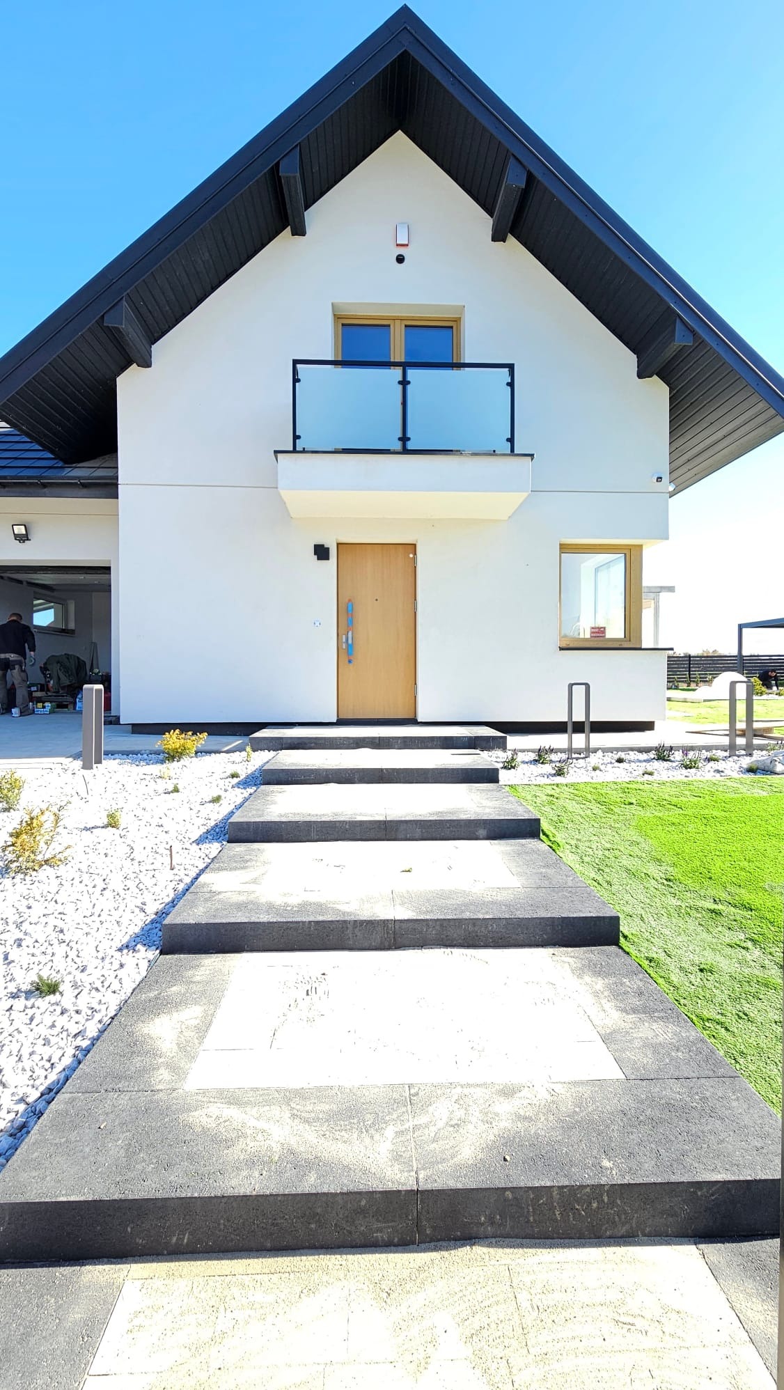 Nowoczesny dom jednorodzinny z wejściem wyłożonym jasnoszarą kostką brukową z ciemnoszarym obramowaniem, otoczony białym żwirem i sztuczną trawą.