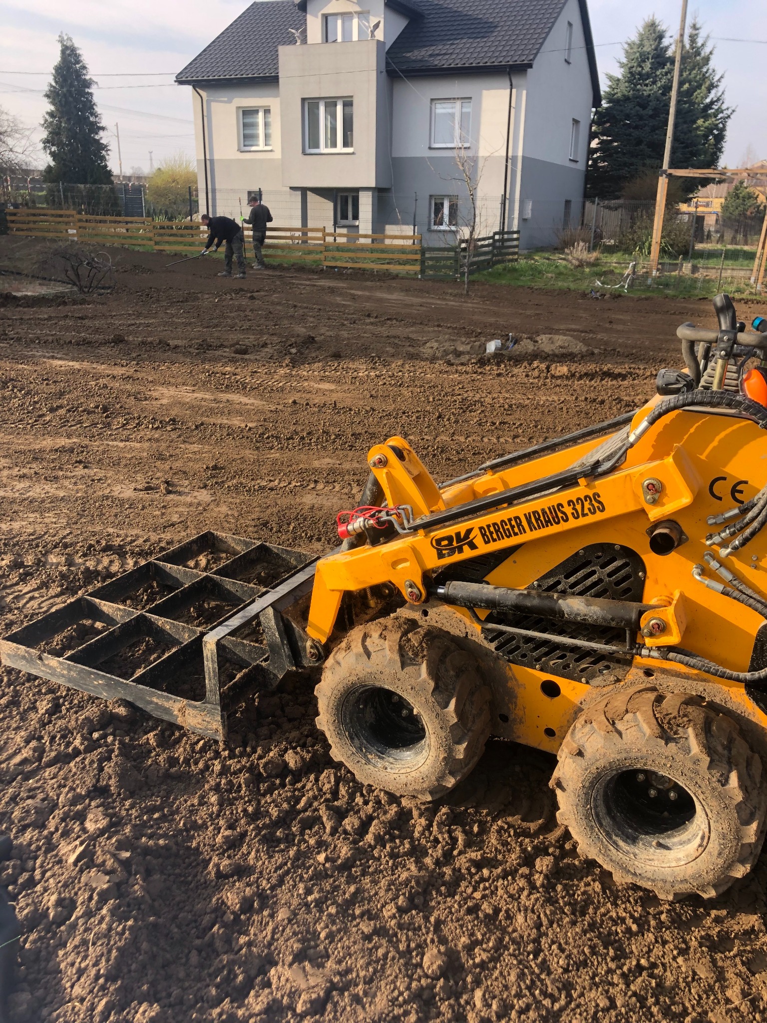 Żółta miniładowarka Berger Kraus 323S z kratą do wyrównywania terenu na placu budowy, w tle dwaj pracownicy z grabiami i ogrodzenie z drewnianych sztachet.