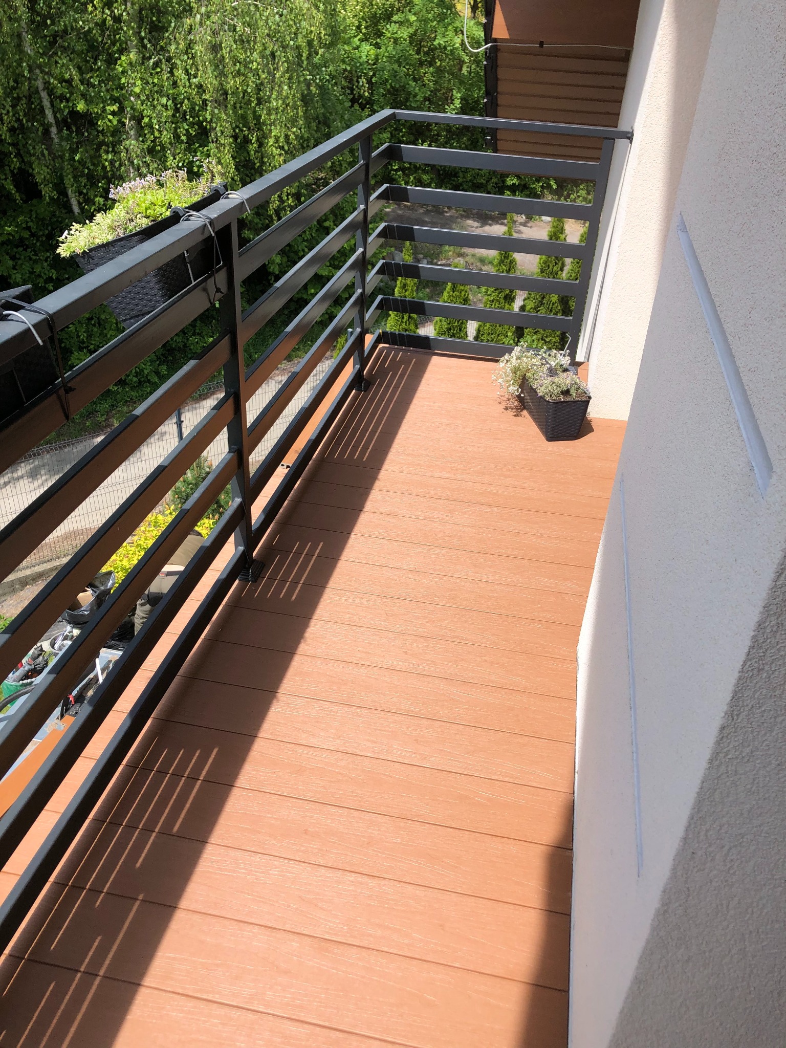 Balkon z brązową deską kompozytową, czarną metalową balustradą i doniczkami z kwiatami, widok z góry.