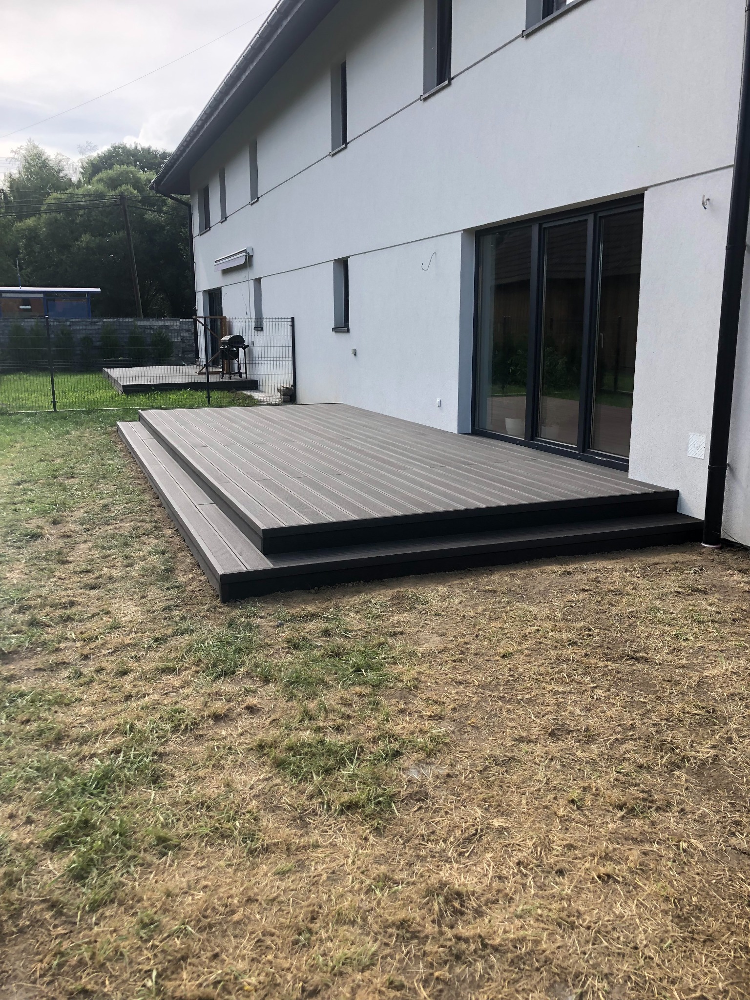 Nowoczesny taras kompozytowy z dwoma stopniami, przylegający do białego budynku z dużymi oknami przesuwnymi, widok z poziomu trawnika.