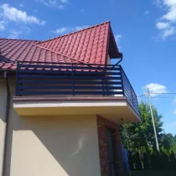 Czarna, pozioma balustrada balkonowa z metalowych profili zamontowana na balkonie domu jednorodzinnego z czerwoną dachówką, widok z dołu na tle błękitnego nieba.