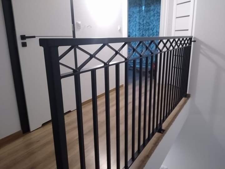 Czarna metalowa balustrada wewnętrzna z geometrycznym wzorem rombów, widok z góry schodów na korytarz z drewnianą podłogą i białymi drzwiami.