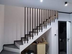 Nowoczesna klatka schodowa z czarnymi pionowymi słupkami balustrady, szare stopnie, białe ściany i czarne obramowanie drzwi.