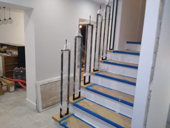 Montaż czarnych, nowoczesnych balustrad schodowych z metalu, zabezpieczonych folią ochronną, na drewnianych schodach z niebieską taśmą malarską.