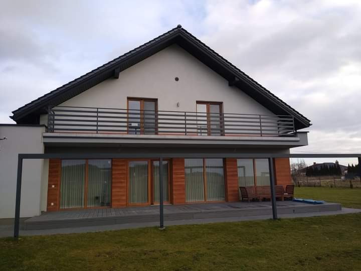 Nowoczesny dom jednorodzinny z szarą, poziomą balustradą balkonową, drewnianą elewacją parteru i zadaszonym tarasem z szarą podłogą.