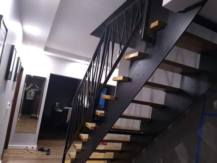 Nowoczesne schody z czarną, metalową balustradą o geometrycznym wzorze i drewnianymi stopniami, widoczne z dołu, w tle lustro z odbiciem osoby.