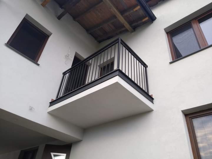 Czarny, metalowy balkon z prostymi barierkami, zamontowany na białej elewacji budynku z drewnianymi oknami, widziany z dołu pod kątem.