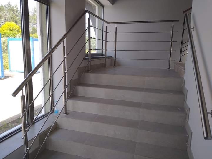 Schody wyłożone szarymi płytkami z balustradą ze stali nierdzewnej z poziomymi prętami, widoczne przez okno drzewa