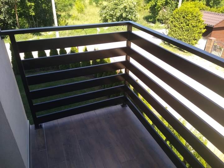 Nowoczesna, czarna balustrada balkonowa z poziomymi szczeblami, widok z góry na drewnopodobną podłogę balkonu i zielone otoczenie.