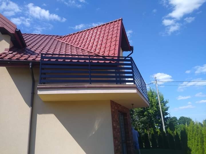 Czarna, pozioma balustrada balkonowa z metalowych profili zamontowana na balkonie domu jednorodzinnego z czerwoną dachówką, widok z dołu na tle błękitnego nieba.