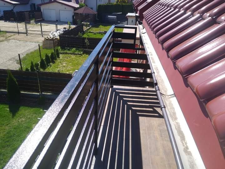 Ciemna, drewniana balustrada balkonowa z widokiem na zadbany ogród i domy w tle, ujęcie z góry.