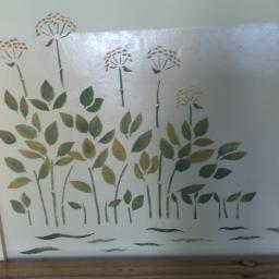 Rem-lux - Ręcznie malowana dekoracja ścienna z motywem roślinnym w odcieniach zieleni i brązu, imitująca łąkę z kwiatami i liśćmi, umieszczona na jasnej ścianie w pokoju.