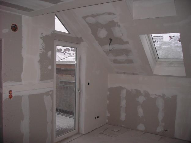 Poddasze w trakcie remontu, ściany i sufit pokryte gipsem, widoczne okno dachowe i drzwi balkonowe z widokiem na zimowy krajobraz.