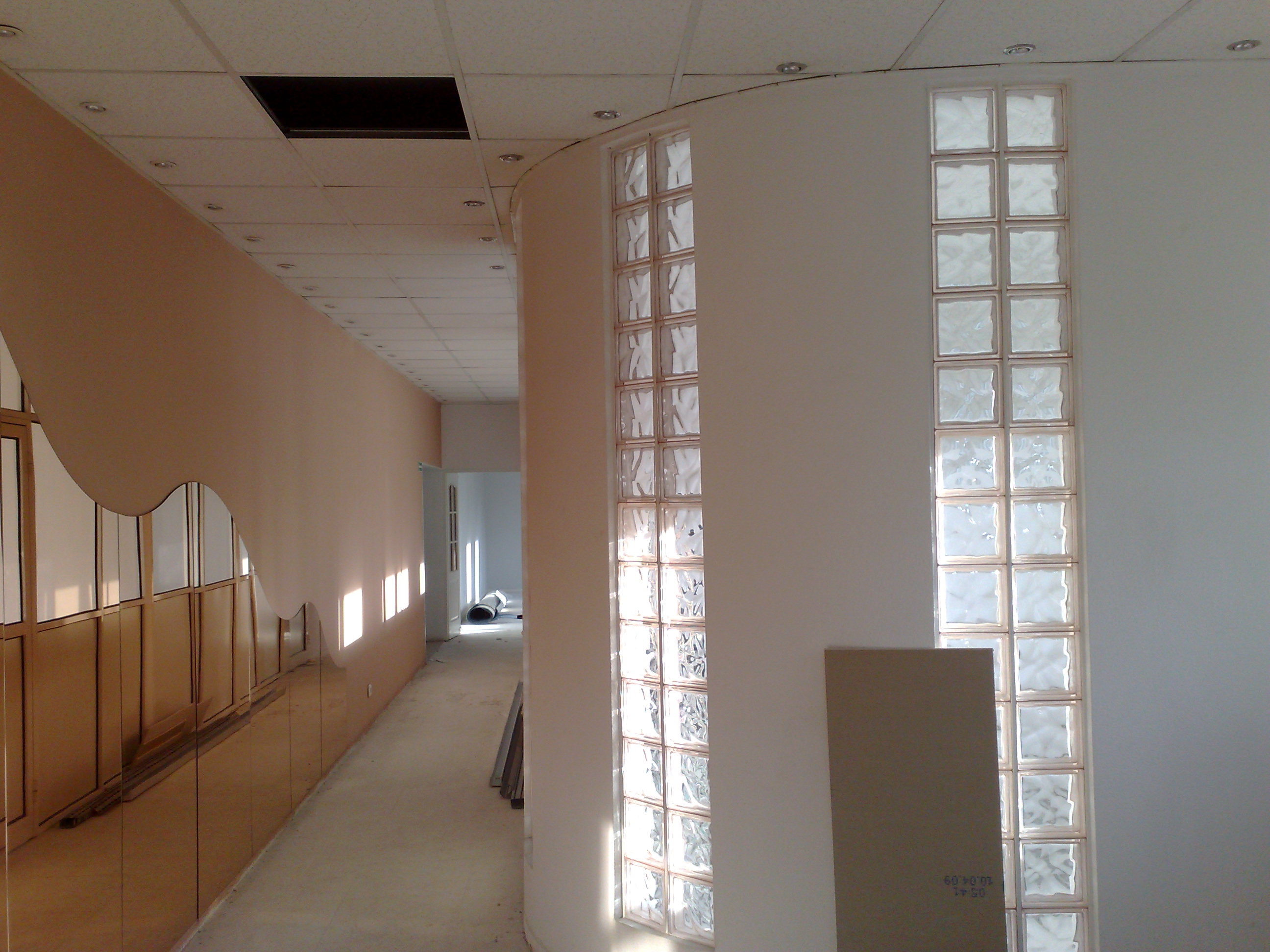 Korytarz w trakcie remontu z oknami z luksferów i falistą ścianą, widoczne materiały budowlane na podłodze.