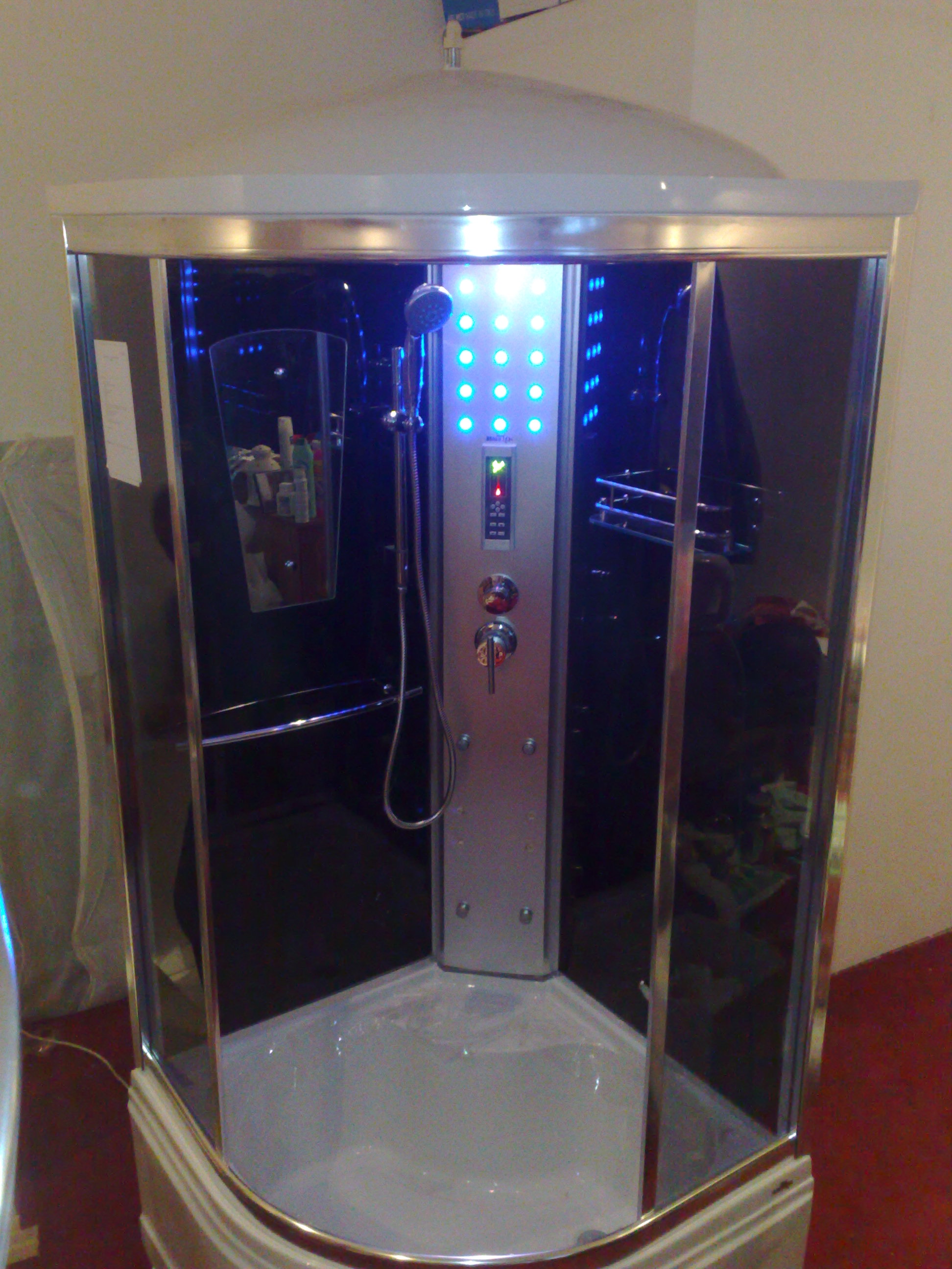 Nowoczesna kabina prysznicowa z hydromasażem, podświetlana niebieskimi diodami, widoczne panele sterowania i dysze, lustro z kosmetykami w tle, chromowane wykończenia.