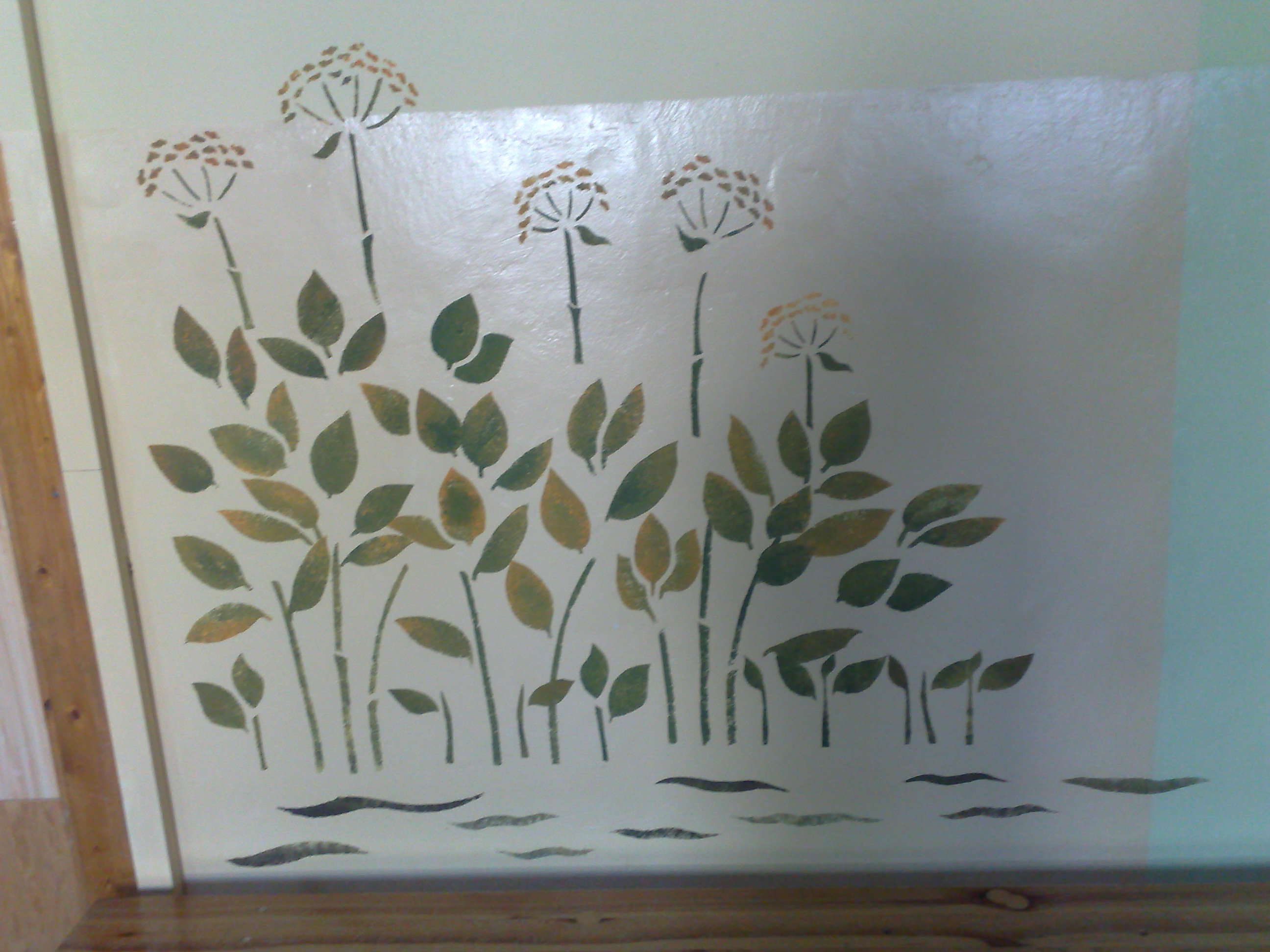 Ręcznie malowana dekoracja ścienna z motywem roślinnym w odcieniach zieleni i brązu, imitująca łąkę z kwiatami i liśćmi, umieszczona na jasnej ścianie w pokoju.