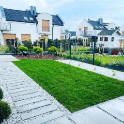 Uporządkowany ogród z równo przystrzyżonym trawnikiem, ścieżką z płyt betonowych otoczoną białym żwirem, kulistymi krzewami po lewej i pracownikiem zamiatającym płytki tarasowe po prawej stronie...