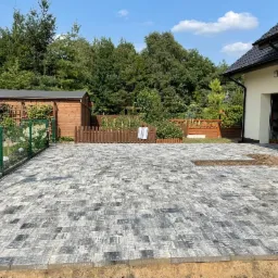 Ułożona nawierzchnia z szarej kostki brukowej na posesji domu jednorodzinnego, widoczny fragment niezakończonej pracy, drewniana altana w tle.