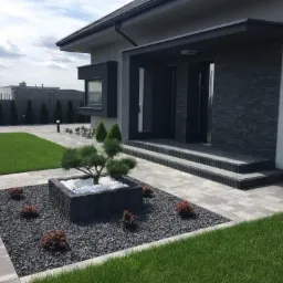 Nowoczesny taras kamienny z szarej kostki brukowej prowadzący do wejścia domu, z centralnie umieszczoną donicą z bonsai i białymi kamieniami, otoczoną czarnym grysem i niskimi krzewami.