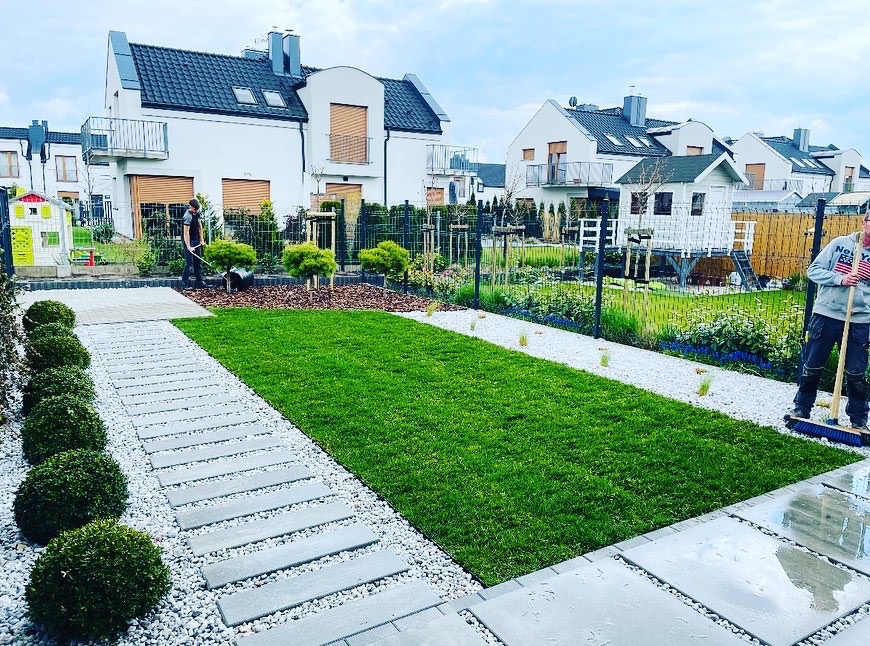 Uporządkowany ogród z równo przystrzyżonym trawnikiem, ścieżką z płyt betonowych otoczoną białym żwirem, kulistymi krzewami po lewej i pracownikiem zamiatającym płytki tarasowe po prawej stronie...