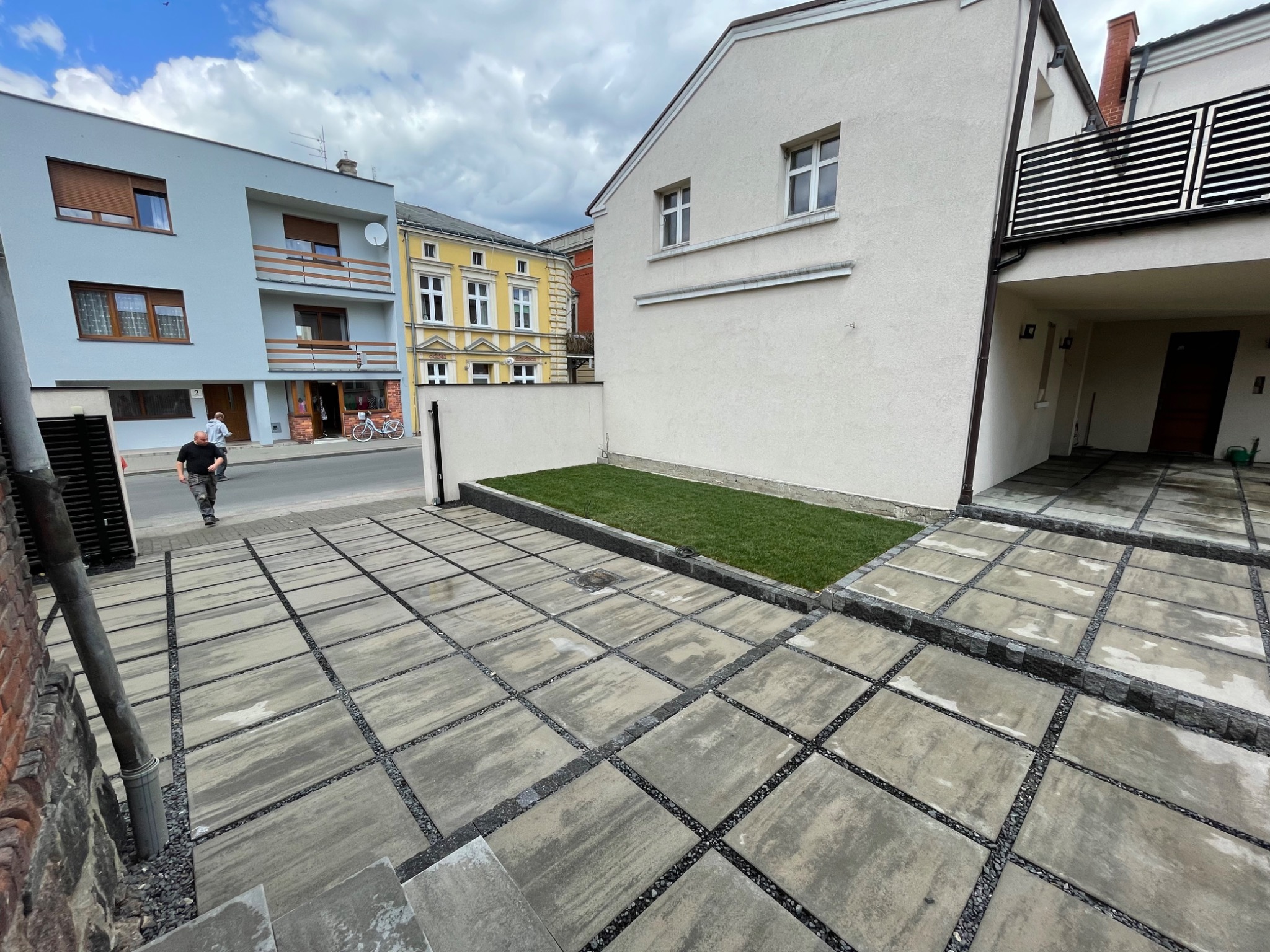 Nowoczesny taras/podjazd z szarej kostki brukowej z czarnymi fugami żwirowymi, przylegający do budynku z małym trawnikiem, widok na ulicę i inne budynki.