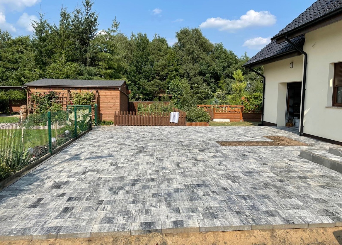 Ułożona nawierzchnia z szarej kostki brukowej na posesji domu jednorodzinnego, widoczny fragment niezakończonej pracy, drewniana altana w tle.