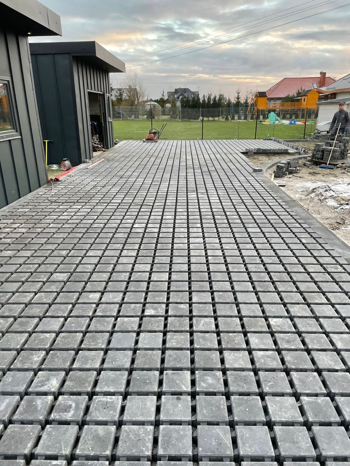 Układanie szarej kostki brukowej na tarasie, widoczna część powierzchni już wyłożona, w tle budynek gospodarczy, trawnik i pracownik układający obrzeża.