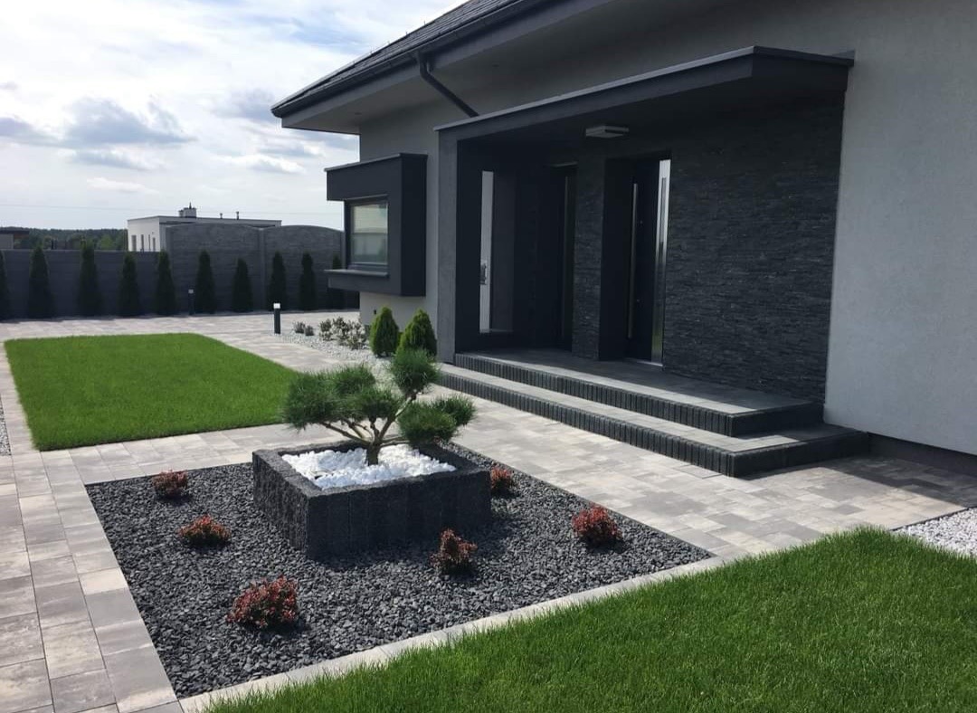 Nowoczesny taras kamienny z szarej kostki brukowej prowadzący do wejścia domu, z centralnie umieszczoną donicą z bonsai i białymi kamieniami, otoczoną czarnym grysem i niskimi krzewami.