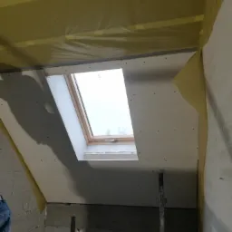 Montaż okna dachowego na poddaszu z izolacją folią paroizolacyjną, widoczne płyty kartonowo-gipsowe i rusztowanie.