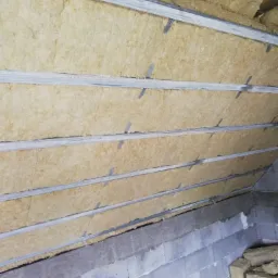 Skośny sufit poddasza z zamontowaną wełną mineralną i metalowymi profilami, widoczne fragmenty drewnianej konstrukcji dachu.