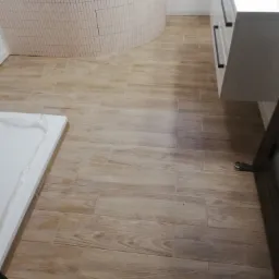 Ułożona podłoga z płytek imitujących drewno w łazience, widoczny fragment wanny z mozaiką i szafka pod umywalką z czarnymi uchwytami.