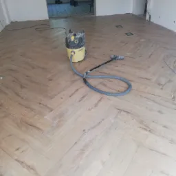 Ułożona drewniana podłoga w jodełkę, z widocznym odkurzaczem przemysłowym Karcher i fragmentami niepomalowanych ścian w tle.