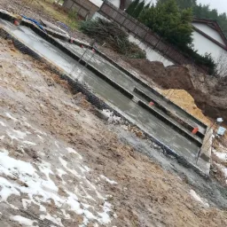 Wylane fundamenty pod budynek jednorodzinny z widocznym zbrojeniem i deskowaniem, teren wokół częściowo pokryty śniegiem.