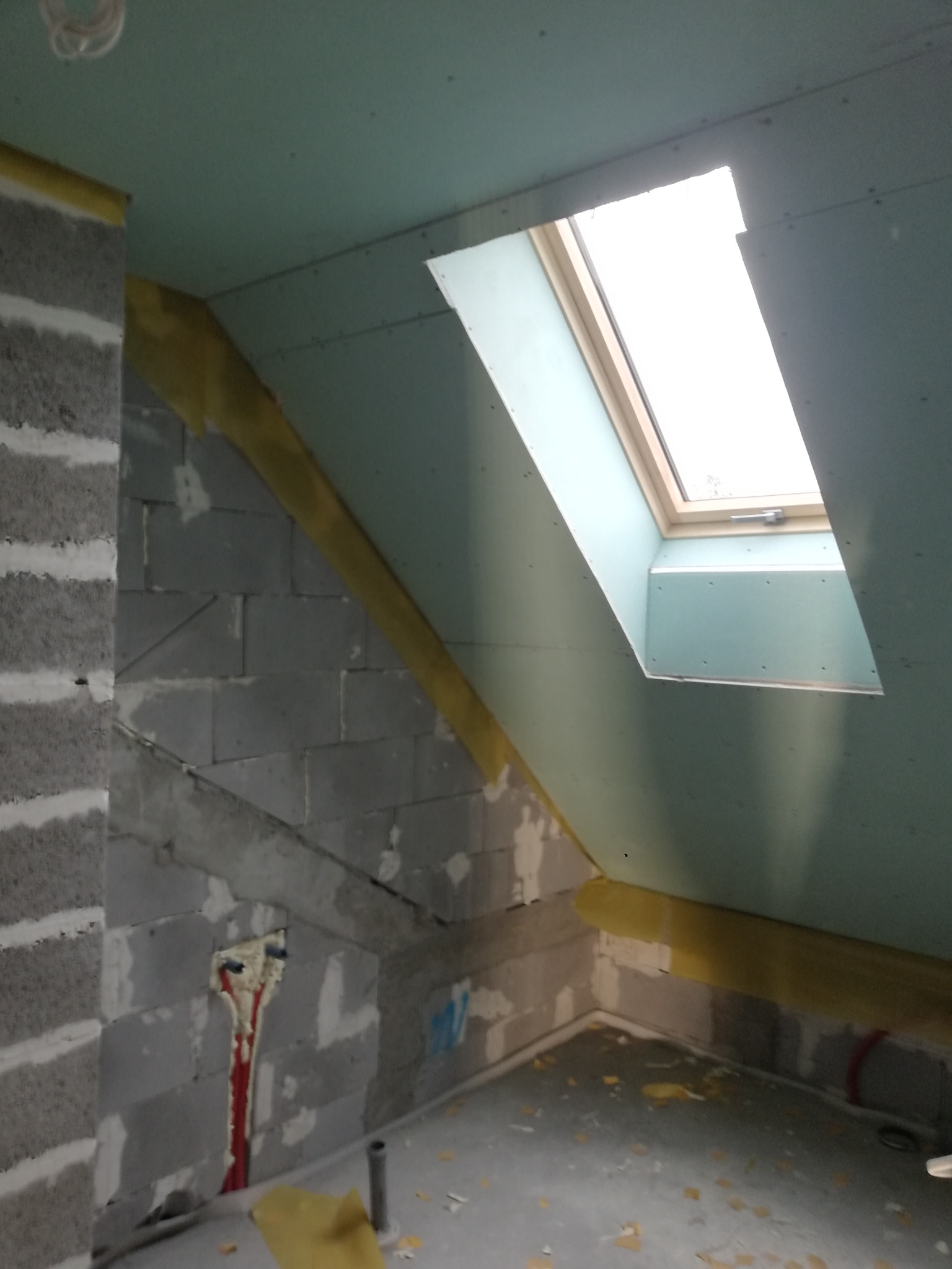 Poddasze w trakcie wykańczania: surowe ściany z betonu komórkowego, częściowo zakryte płytami kartonowo-gipsowymi w kolorze jasnoniebieskim, okno dachowe z drewnianą ramą oraz widoczne elementy...