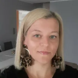 Portret blondynki z dużymi, okrągłymi kolczykami w panterkę, stojącej w jasnym, minimalistycznym wnętrzu.