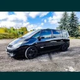 Czarny, błyszczący van Renault Espace z czarnymi felgami stoi na żwirowym podłożu; w tle drzewa i błękitne niebo z chmurami.