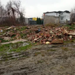 Teren po rozbiórce budynku z gruzem ceglanym, błotnistym podłożem i fragmentami zieleni, w tle budynek w stanie surowym.