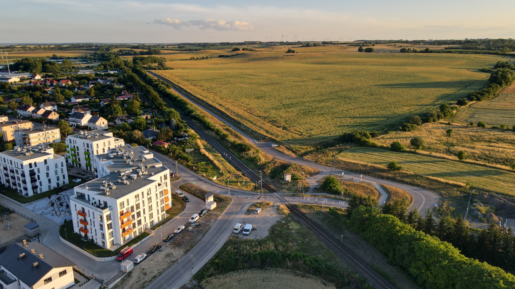 Zdjęcie z lotu ptaka przedstawiające nowoczesne, białe budynki mieszkalne z kolorowymi balkonami na skraju miasta, przylegające do pól uprawnych, z widoczną linią kolejową i drogą.