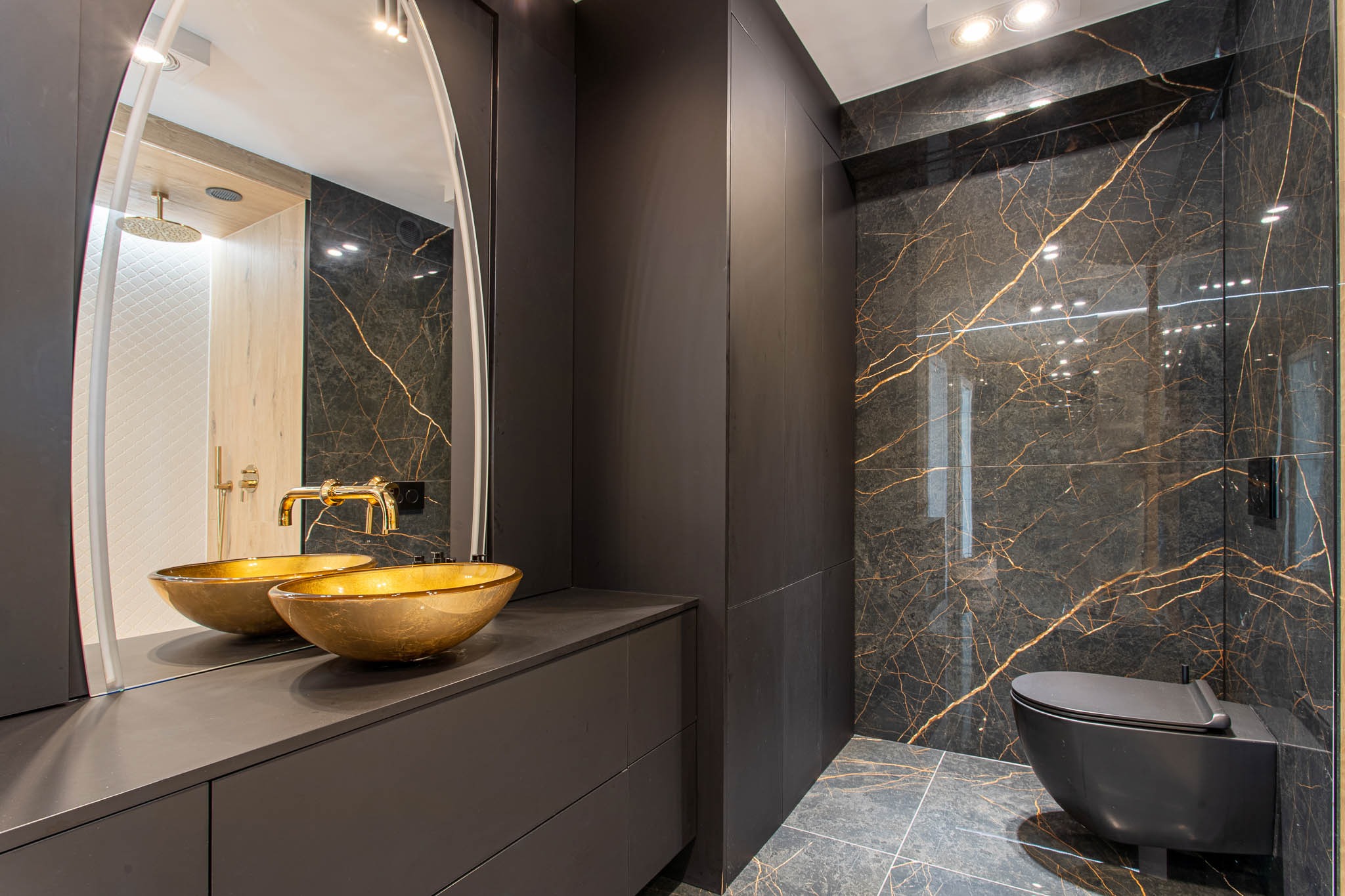 Nowoczesna łazienka z czarnymi płytkami imitującymi marmur ze złotymi żyłami, złotymi umywalkami w kształcie misy i czarną armaturą, widoczna w lustrze kabina prysznicowa z białymi płytkami.