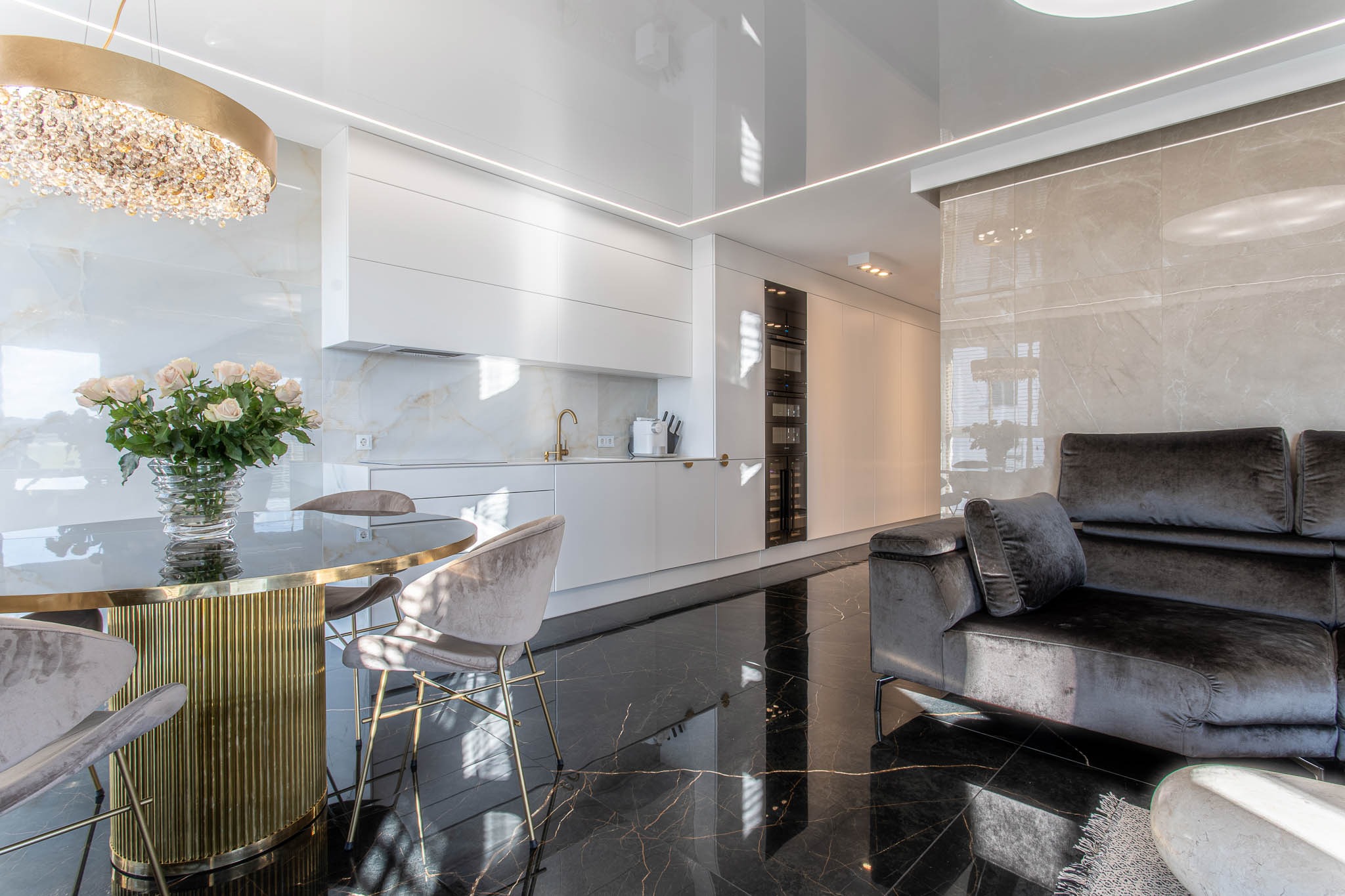 Nowoczesne wnętrze: błyszcząca czarna podłoga, biała zabudowa kuchenna z złotą armaturą, okrągły stół ze złotą podstawą i bukietem róż, szary narożnik z poduszkami, kryształowy żyrandol i marmurowe...
