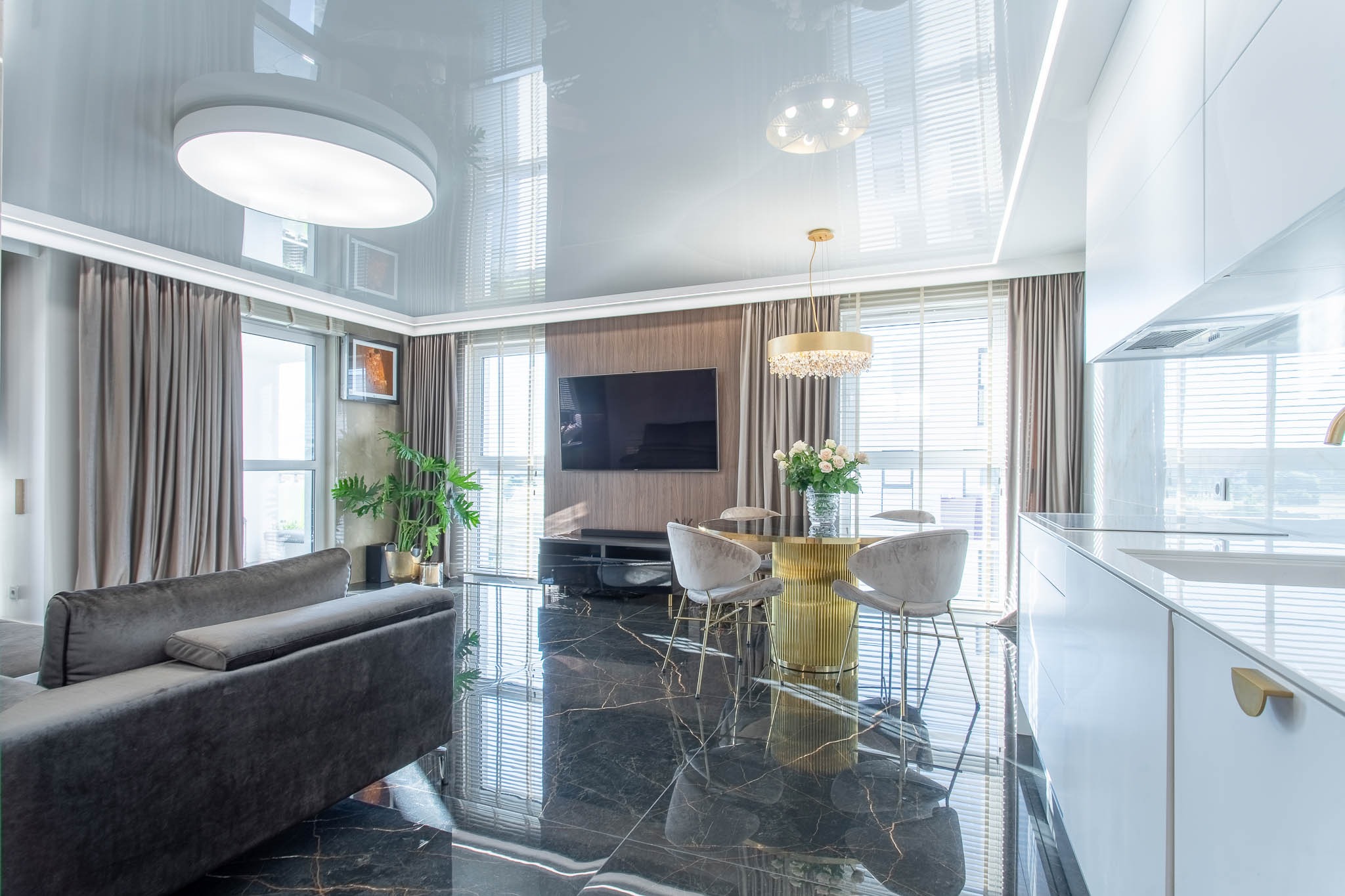 Nowoczesne wnętrze z czarną marmurową podłogą, szarym narożnikiem, złotym stołem jadalnym i białymi meblami kuchennymi pod błyszczącym sufitem.