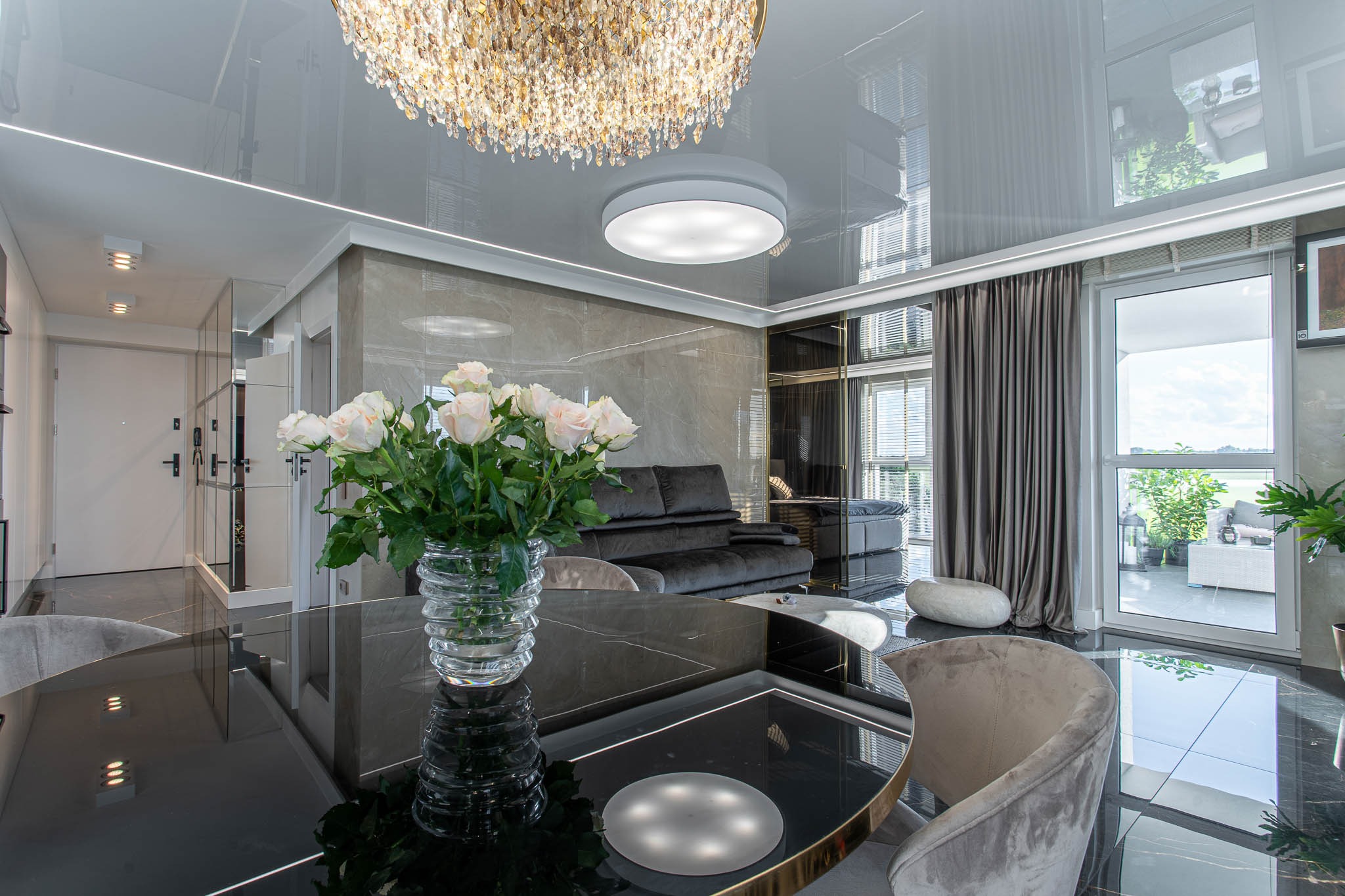 Nowoczesny salon z czarnym, błyszczącym stołem, bukietem białych róż w szklanym wazonie, szarą sofą w tle i dekoracyjnym żyrandolem kryształowym pod błyszczącym sufitem.