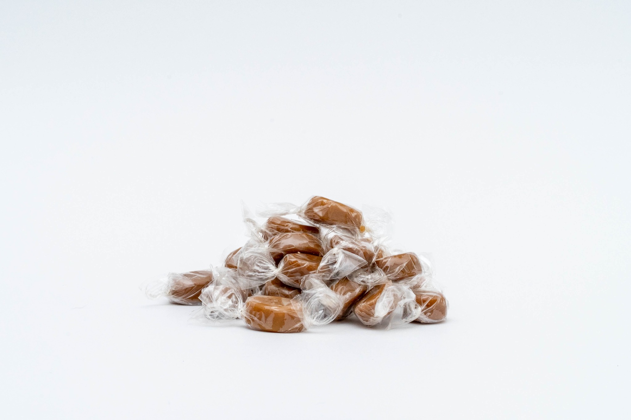 Stos brązowych, karmelowych cukierków owiniętych w przezroczysty celofan na białym tle, oświetlonych studyjnie.